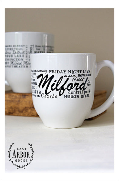 Two white coffee mugs with one showing the front design of words wrapped around the mug in black screen print and in different fonts and sizes. In the middle is the word "Milford" for the town in Michigan. The other words represent places and activities in the area. The second cup is in the background on a book. 