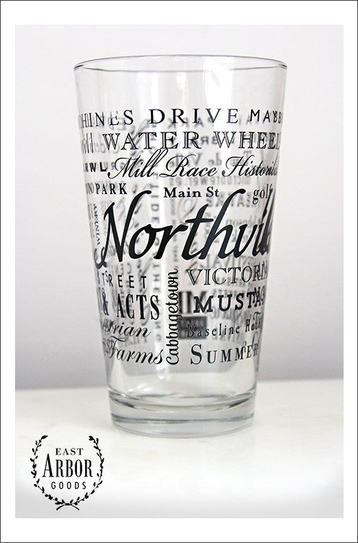 Pint Glass featuring Northville, Michigan with the word Northville in large script in the middle and words of places and activities in different fonts and sizes surrounding. All of the words in black screen print. 