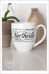 White ceramic coffee mug featuring Northville, Michigan with places and activities in different fonts and sizes in black screen print wrapped around the center of the mug. Placed on a white counter with a brown bag of coffee and plant in the background.  