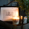 Lit candle in a glass container with a black screen print original sketch of buildings in downtown Northville. Framed sketch behind the candle with both sitting atop a small shelf. 