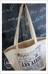 View from above of hanging large natural colored canvas tote bag featuring Ann Arbor, Michigan. Words in different fonts and sizes showcase local places and activities.