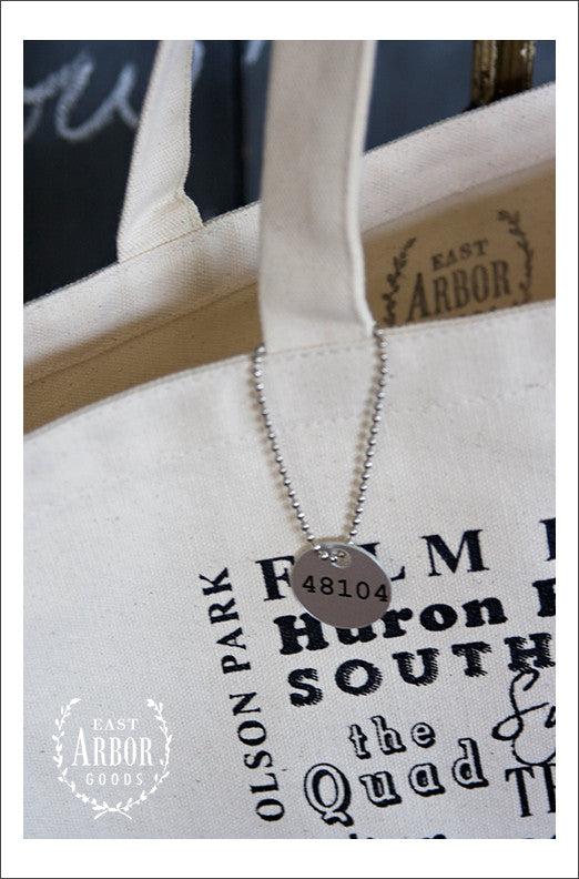 Close up view of small metal tag with Ann Arbor zip code “48104” hanging from one of the tote bag handles.
