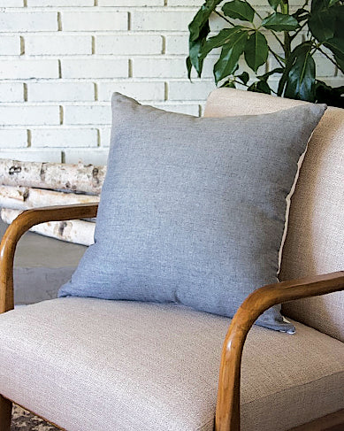 Close up of large pillow on chair in front of white brick wall. Pillow is a blue-gray linen on one side and white linen on the back. 