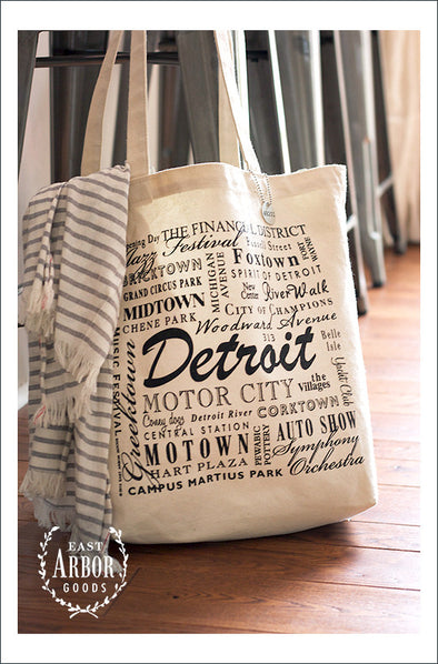 Large natural colored canvas tote bag hanging from a chair with a shawl draped over the side. The bag has words in different fonts and sizes featuring places and activities connected to Detroit, Michigan.