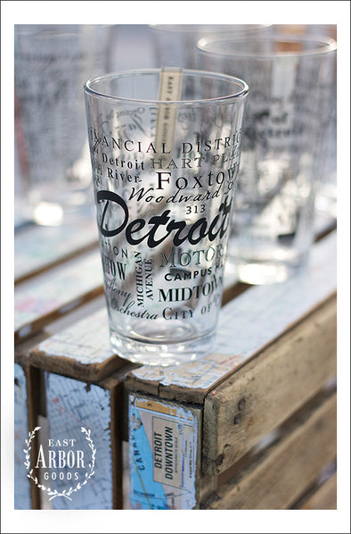A group of pint glasses on a wood crate with one main one in focus featuring Detroit, Michigan with highlights from the town in black screen print.