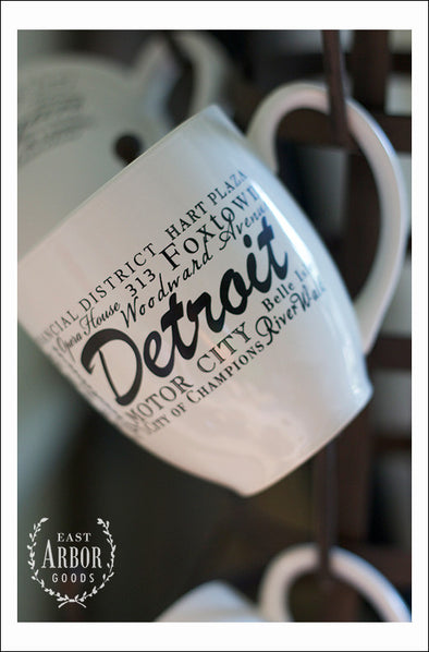 White ceramic coffee mugs hanging on a coffee mug stand focusing on one up close featuring Detroit, Michigan with places and activities featured in different fonts in black screen print. 