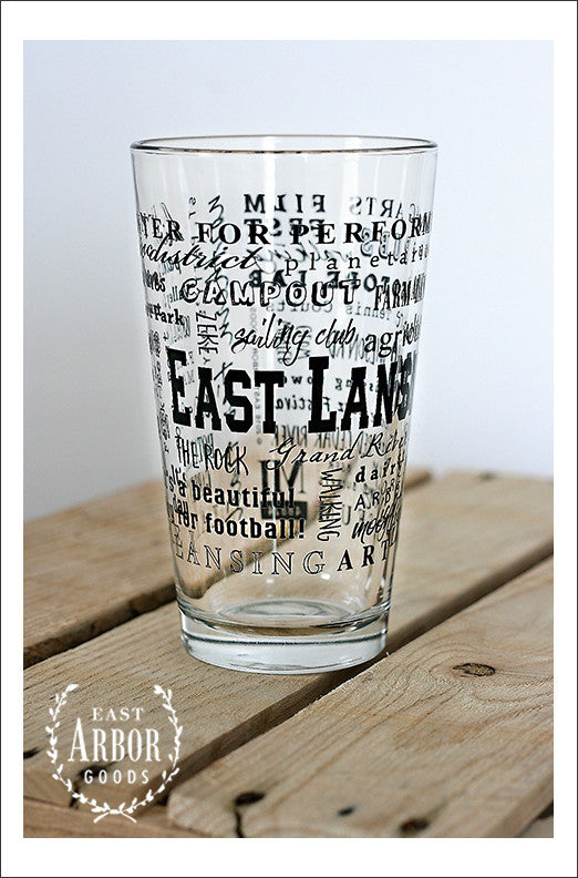 Pint Glass on wood crate. Glass features East Lansing, Michigan with highlights from the town in black screen print.
