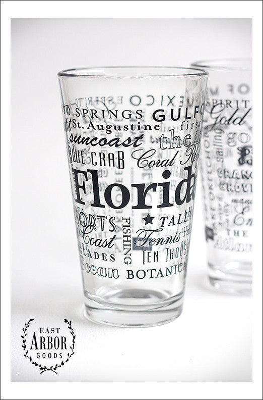 Two pint glasses with one in the center and main focus to show the black screen print details of words in different fonts and sizes featuring highlights of places and activities in Florida. 