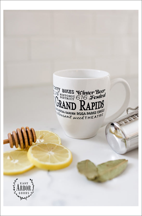 White ceramic coffee mug with lemon slices, honey stick, mint leaves, and tea strainer.  Mug features Grand Rapids, Michigan with black screen-print words in different fonts and sizes.