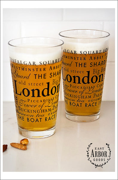 Two pint glasses next to each other filled with beer. Glasses have words in different fonts and sizes in black screen print around the theme of London, England. 