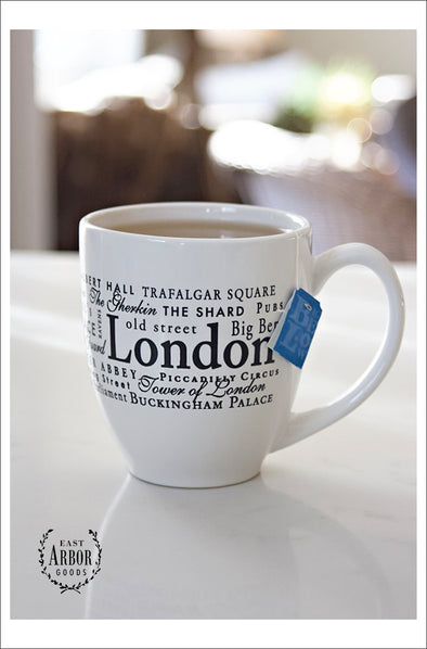 White ceramic mug filled with tea and a blue tea bag tag hanging over on a white countertop. Mug contains words in different fonts and sizes around the theme of London, England.