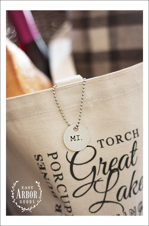 Close up of circular metal tag hanging from one of the handles of a natural cream colored canvas tote bag. The metal tag is stamped with the Michigan state abbreviation "MI."