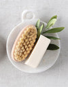 White marble dish with small handle holding a dish brush, stick of butter, and with sage leaves. Dish on top of white canvas tablecloth. 
