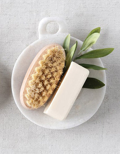 White marble dish with small handle holding a dish brush, stick of butter, and with sage leaves. Dish on top of white canvas tablecloth. 