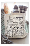 Large natural colored canvas tote bag placed on a chair and filled with a baguette and gray and white blanket. The design on the bag is made up of words in black ink and in different fonts and sizes around the theme of the state of Michigan. In the center in large script is the word "Michigan."