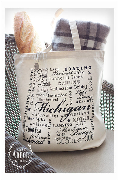 Large natural colored canvas tote bag placed on a chair and filled with a baguette and gray and white blanket. The design on the bag is made up of words in black ink and in different fonts and sizes around the theme of the state of Michigan. In the center in large script is the word "Michigan."