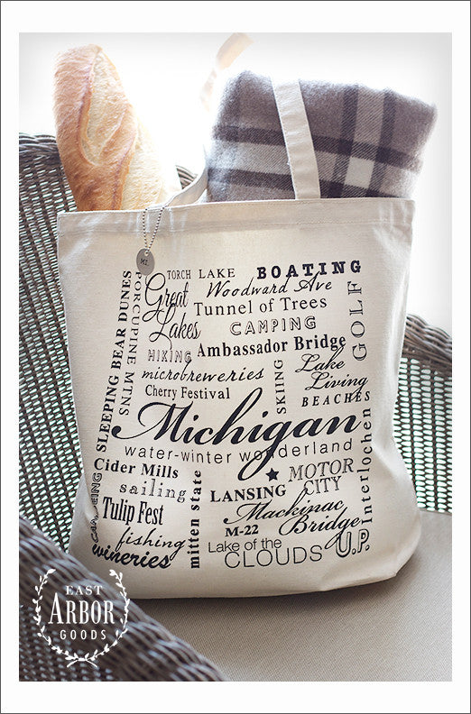 Large natural colored canvas tote bag placed on a chair and filled with a baguette and gray and white blanket. The design on the bag is made up of words in black ink and in different fonts and sizes around the theme of the state of Michigan. In the center in large script is the word "Michigan."
