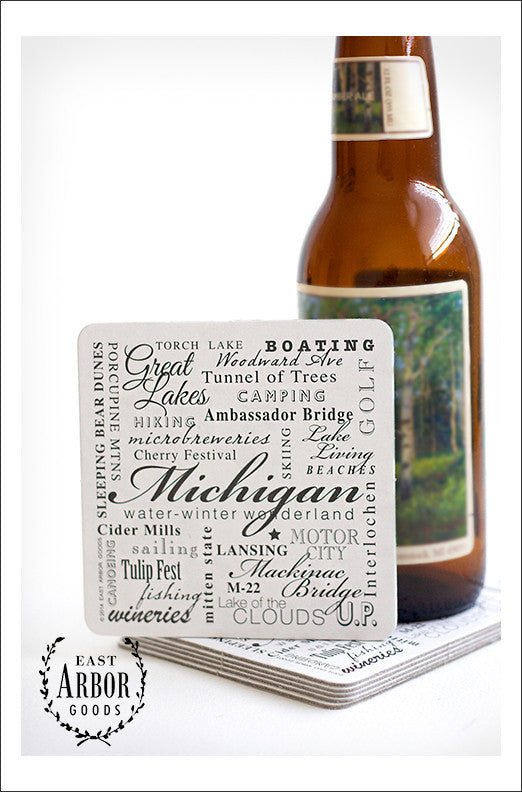 Stack of drink coasters with a glass beer bottle on top and one of the coasters leaning against the bottle to show the coaster design. The design is made up of words in different fonts and sizes around the theme of the state of Michigan.