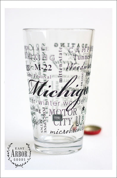 Close up of glass pint glass with design of multiple words around the glass. Words are in the theme of the state of Michigan including different places and activities connected to the state. Small red bottle cap behind the glass. 