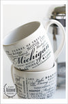 Two white coffee mugs with one stacked on top of the other -- one facing forward, the other with the back showing. The design on the mugs is made up of words in black screen print wrapped around in different fonts and sizes in the theme of the state of Michigan.