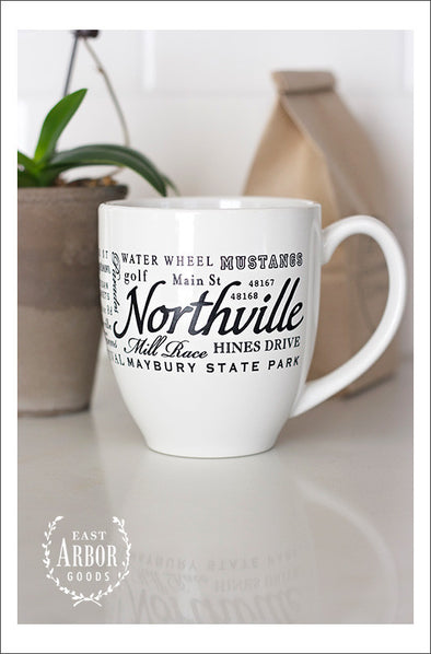 White ceramic coffee mug featuring Northville, Michigan with places and activities in different fonts and sizes in black screen print wrapped around the center of the mug. Placed on a white counter with a brown bag of coffee and plant in the background.  