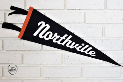 Black felt pennant with orange accent detail and the town "Northville" in large white script. Pennant hanging on a white brick background. 