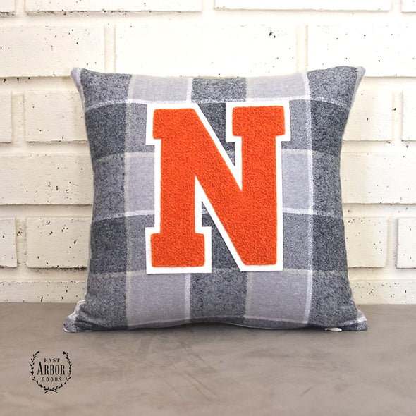 Gray and white large checked accent pillow with a large orange "N" varsity letter sitting on a cement countertop and leaning against a white brick background. 