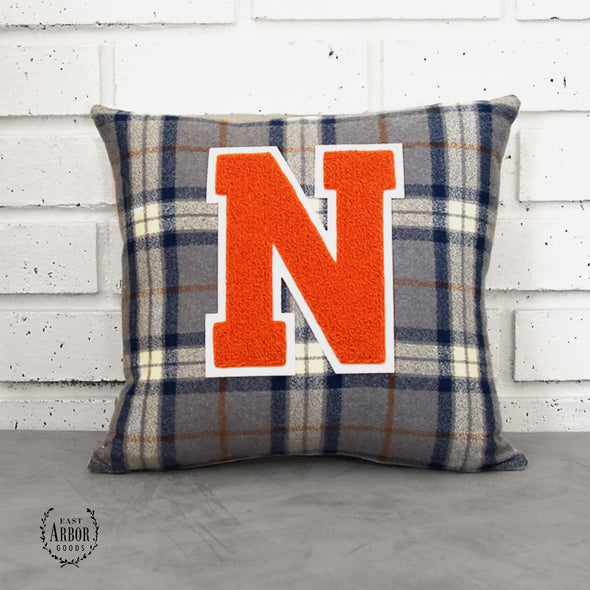 Dark navy blue, cream, and gold large checked accent pillow with a large orange "N" varsity letter sitting on a cement countertop and leaning against a white brick background.