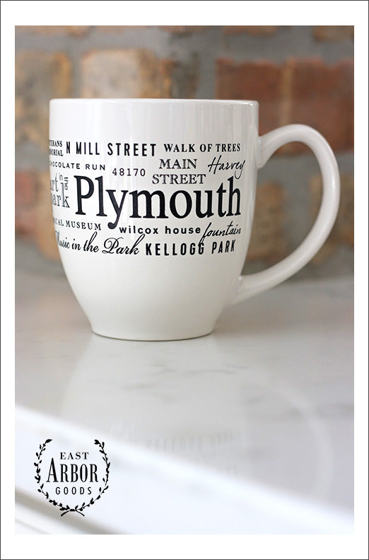 White ceramic coffee mug featuring Plymouth, Michigan with black screen print design of the name "Plymouth" in large letters in the middle and surrounded with words of places and activities in different fonts surrounding it and wrapped around the center of the mug.  Mug on a white countertop and red brick background.