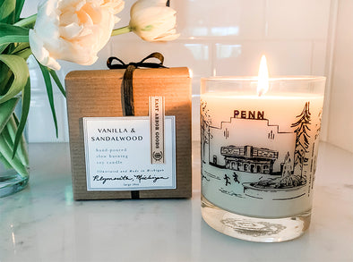 Lit cream colored candle in a glass jar with a hand drawn image of the Penn Theatre in Plymouth, Michigan. Next to lit candle is the brown gift box it comes in with white square label including the Vanilla & Sandalwood name and additional details. Both on a white kitchen countertop and part of white flowers in a vase on the left side. 