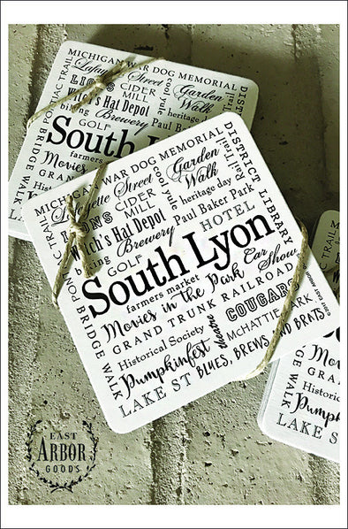 Three sets of rounded square drink coasters shown featuring South Lyon, Michigan in a black text design on cream background. Each set comes with 6 thick cardstock coasters wrapped in decorative twine. Sets laid out on gray brick.