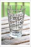 Left side view of Pint Glass featuring South Lyon, Michigan with highlights from the town in black screen print. On an outdoor wood plank table with green grass in the background.