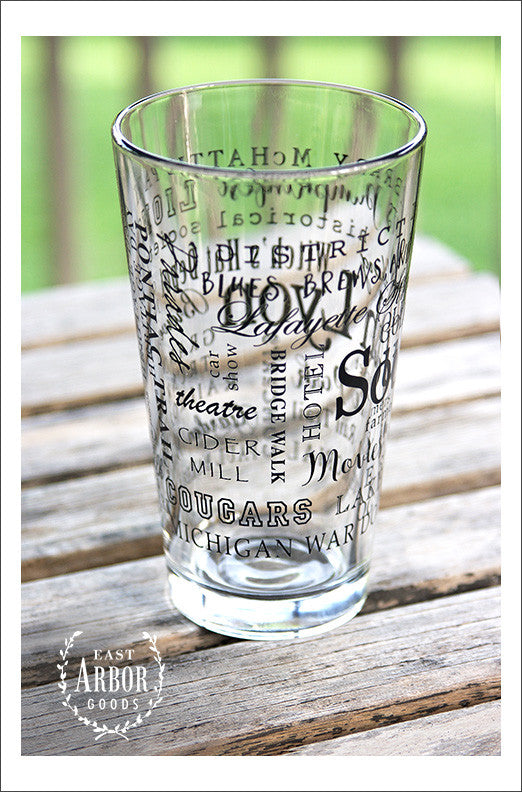 Left side view of Pint Glass featuring South Lyon, Michigan with highlights from the town in black screen print. On an outdoor wood plank table with green grass in the background.