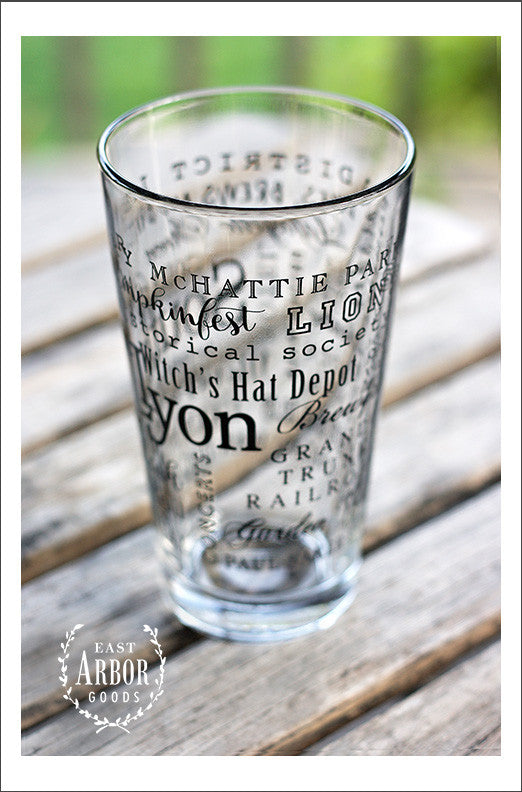 Right side view of Pint Glass featuring South Lyon, Michigan with highlights from the town in black screen print. On an outdoor wood plank table with green grass in the background.
