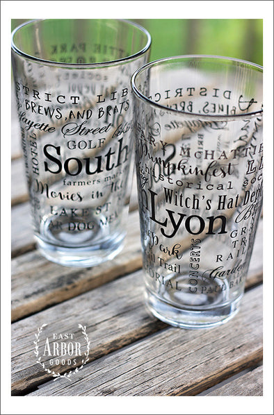 Two pint glasses next to each other on a wooden table and outdoor green grass background. Glass design is made up of words in black screen print and different fonts and sizes with the name "South Lyon" the largest and centered and the other words wrapping around the glass.