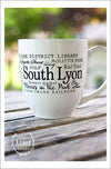 Close up of large white ceramic mug on an outdoor wood table with trees in background. Design on mug wraps around the middle of the mug with words in different sizes and fonts around the main words "South Lyon." Each word related to the town.