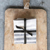 Set of three folded cream and black striped cotton kitchen towels wrapped with a black ribbon and placed in the middle of a rustic cutting board with handle.