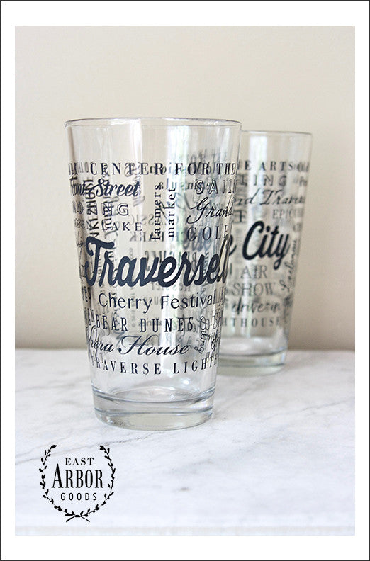 Two clear pint glasses next to each other on a white marble countertop and against a white wall. Glass design is made up of words in black screen print in different fonts and sizes with the words "Traverse City" the largest and centered and the other words wrapping around the glasses.