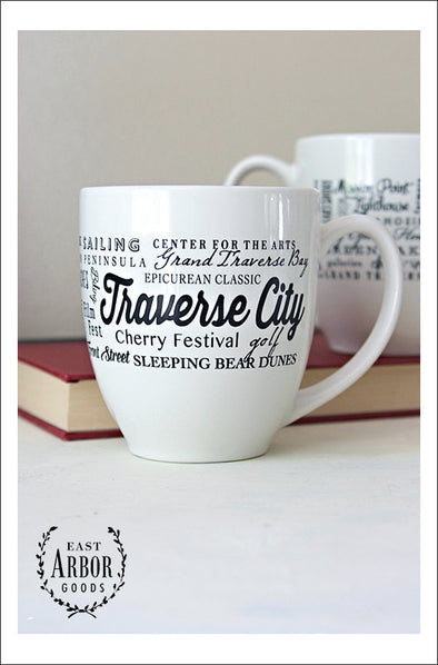Two white ceramic mugs with one centered in the image showcasing the black screen print design of words in different fonts and sizes. Center of design are the words "Traverse City" with different places and activities from the area wrapping around the mug. Second mug is on a book in the background showing the back of the mug and design. 